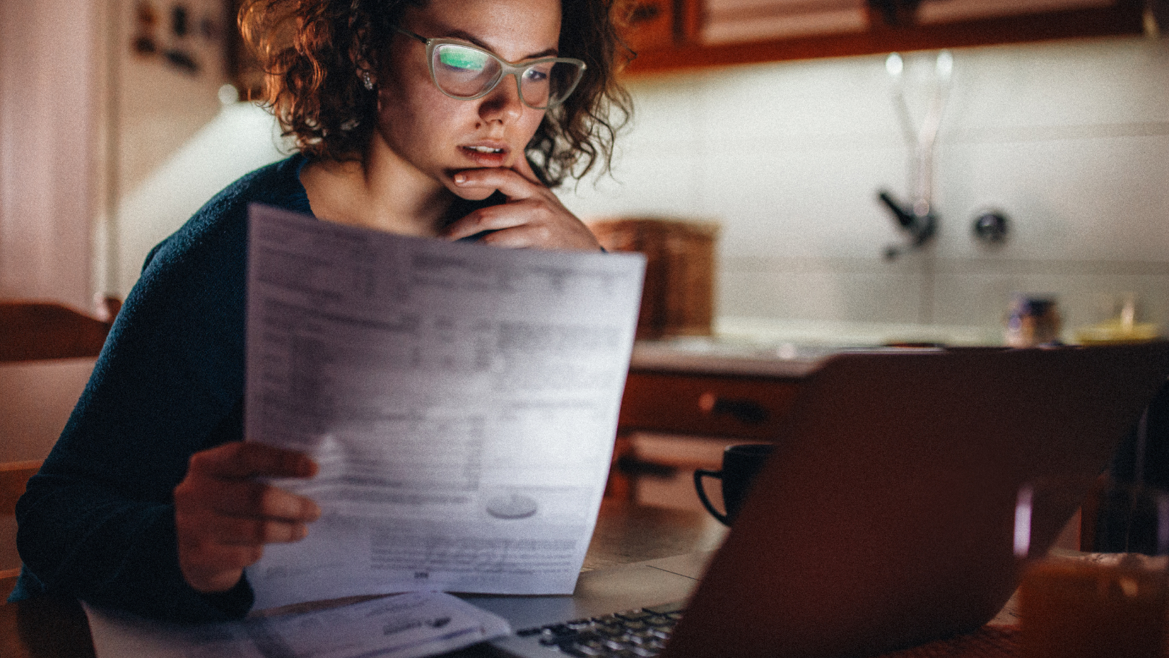 A focused young woman with curly hair and glasses intently reviews financial documents and papers while using her laptop in a dimly lit home kitchen, possibly budgeting or working late.