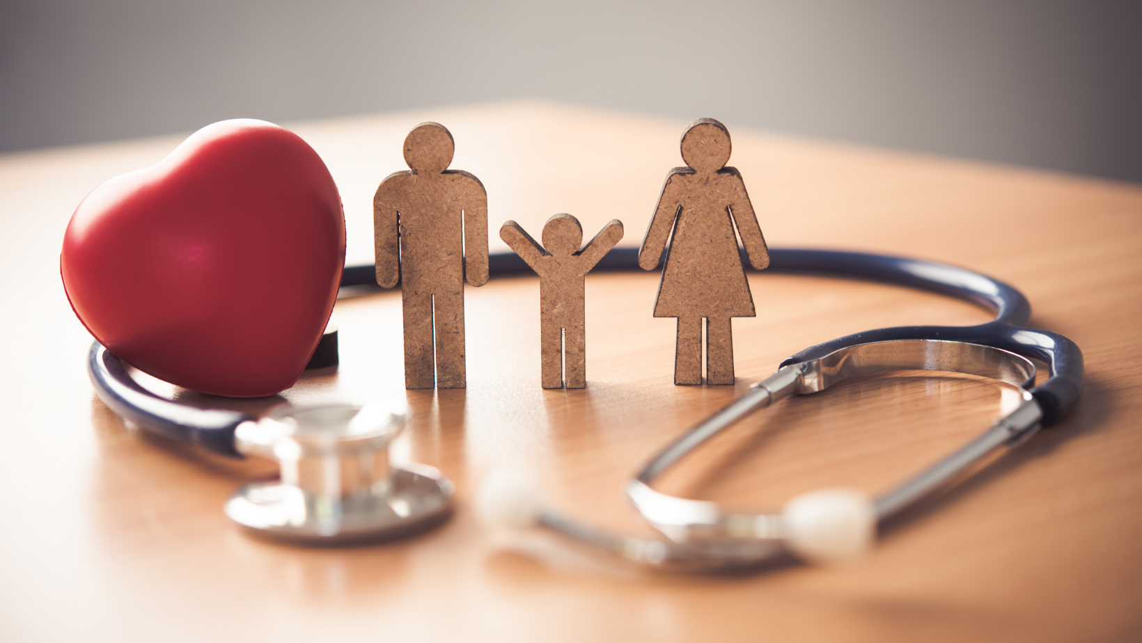 A red heart, wooden family cutouts (father, mother, child), and a blue stethoscope on a light wooden table, symbolizing comprehensive family health and medical care.