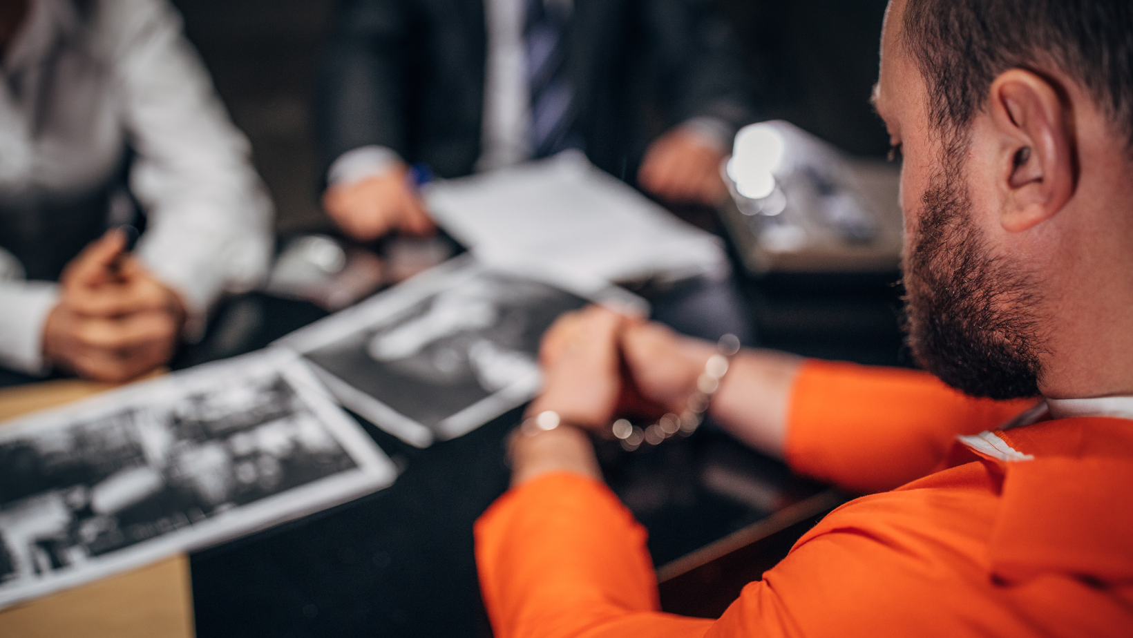 Handcuffed man in an orange jumpsuit sits at a table, being interrogated by blurred law enforcement officers with legal documents and evidence present.