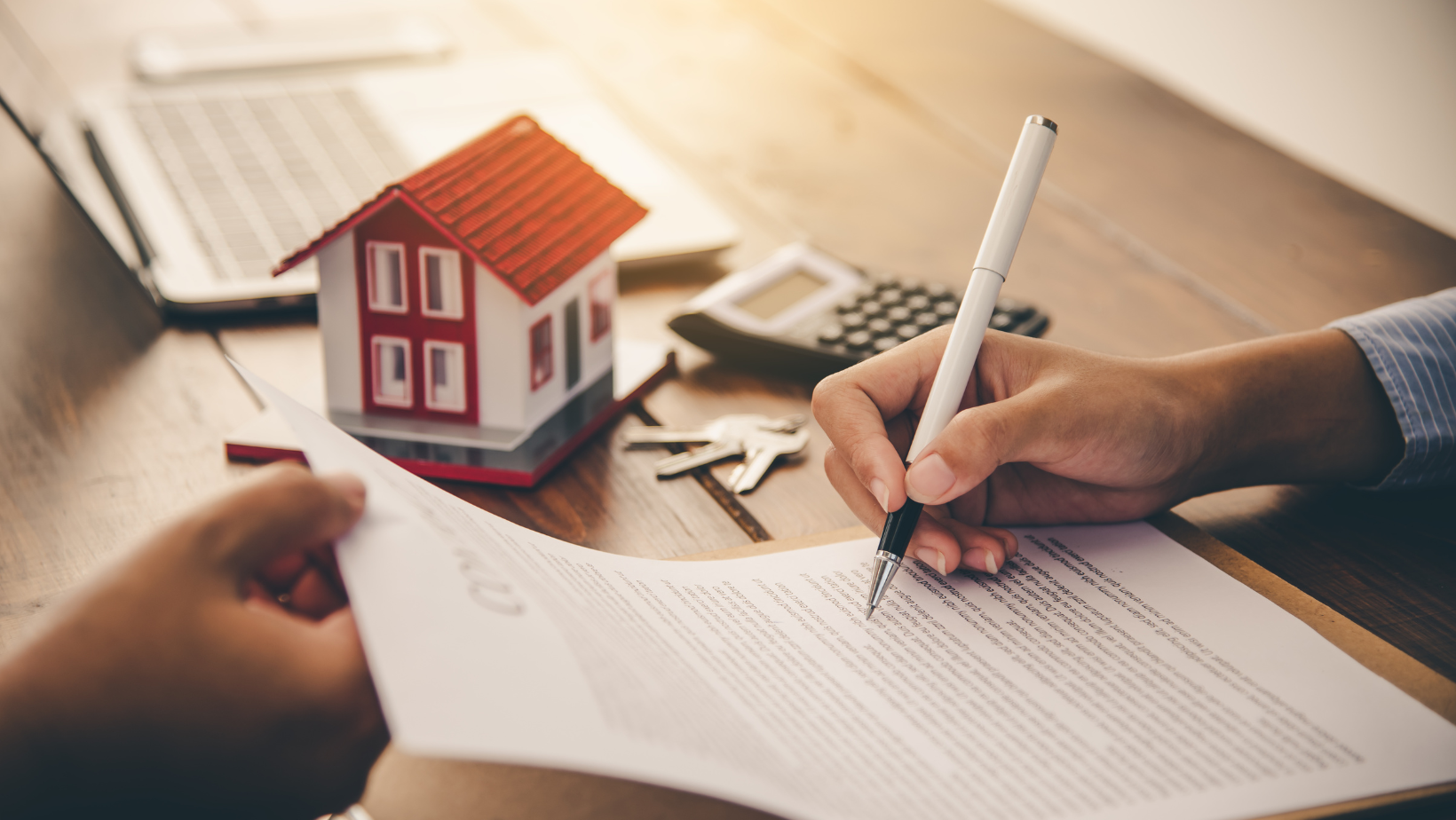 Person signing a real estate contract with a model house, calculator, and keys on the table, representing property purchase or mortgage agreement.