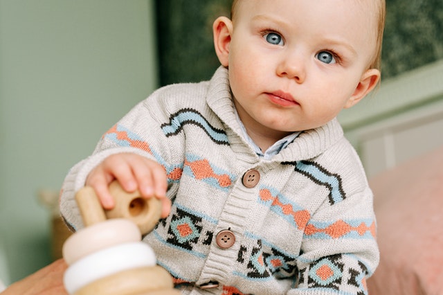Adorable baby boy with bright blue eyes wearing a cozy patterned knit cardigan, gently playing with a natural wooden stacking toy. The baby is focused on the toy.