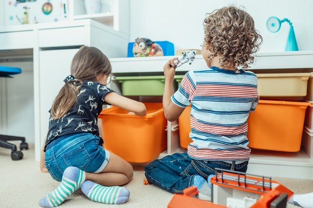 Two young children, a boy and a girl, are playing with toys and organizing items in colorful storage bins on the floor of a brightly lit playroom.