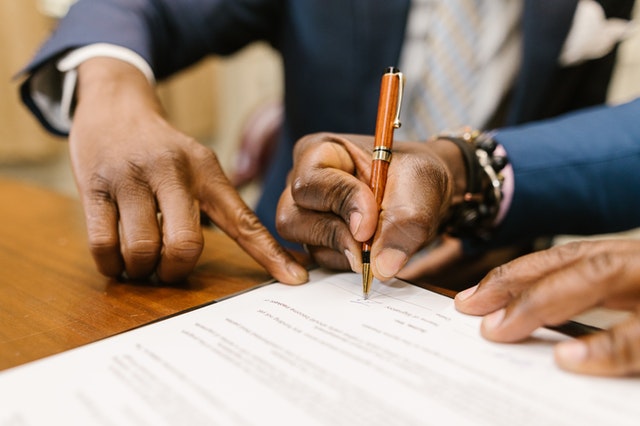 Close-up of two hands, one pointing to a line on a legal document while the other hand signs with a pen, representing a business agreement or contract signing.