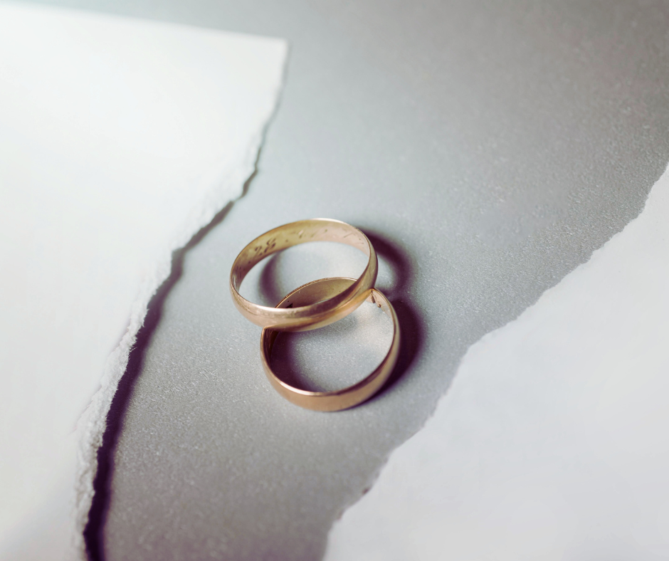 Pair of classic gold wedding bands, one inscribed, on a textured gray surface framed by torn white paper. Evokes timeless love and marital commitment.